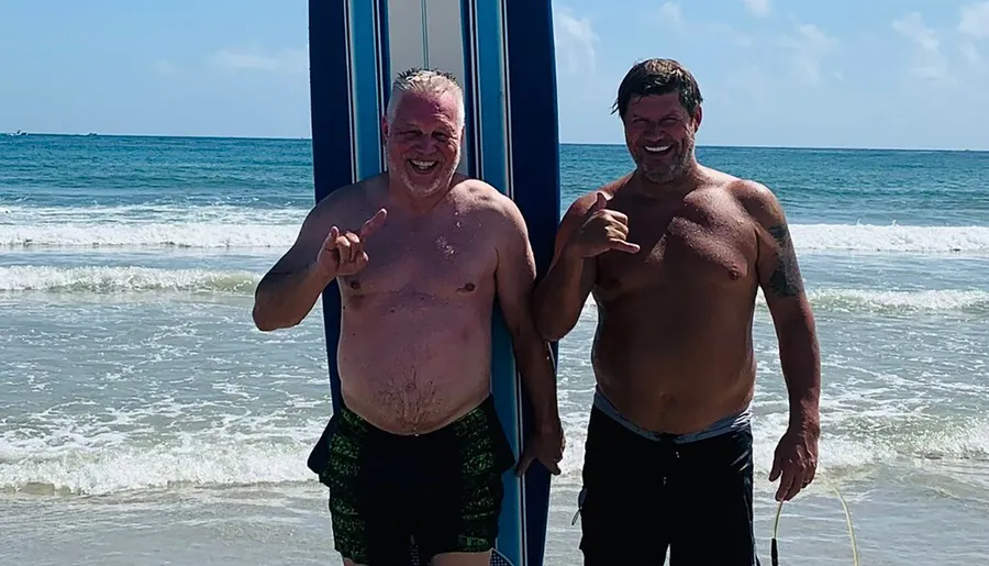 Two men are standing on the beach smiling and gesturing hang loose signs with their hands.