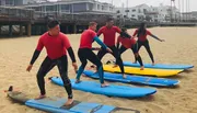 A group of people in wetsuits is practicing surfing stances on surfboards placed on the beach.