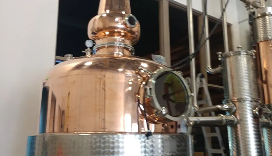 The image shows a copper distillation apparatus, likely used in the production of spirits, with a manway and various pipes and pressure gauges attached.