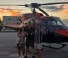 Three people are posing with thumbs up in front of an orange and black helicopter at sunset