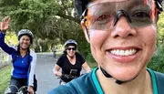 Three people are enjoying a bicycle ride on a scenic path, with the person in the foreground taking a selfie and the other two waving and smiling behind her.