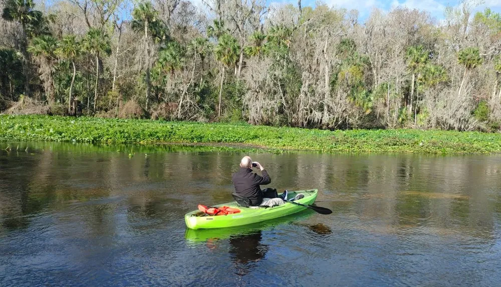 A person in a green kayak is photographing or observing the natural river surroundings