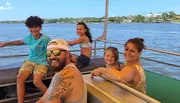 A smiling family of five is enjoying a sunny boat ride with a scenic water view in the background.