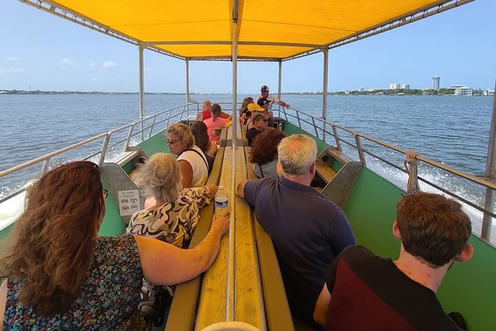 People are seated on a covered boat enjoying a ride on a sunny day with views of the water and buildings in the distance