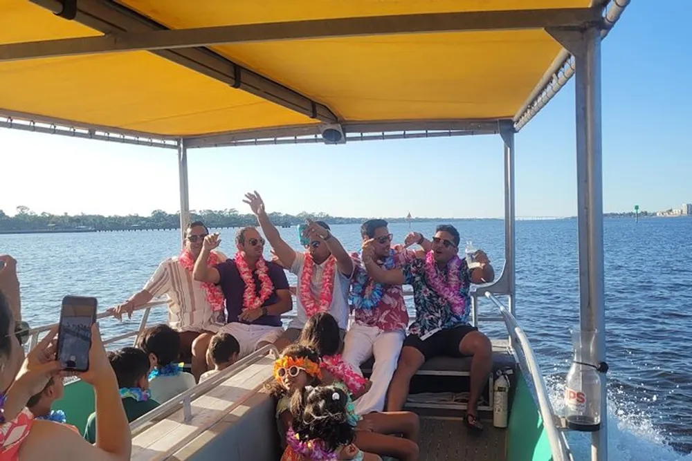 A group of people in colorful attire is enjoying a festive boat ride on a sunny day with some passengers taking photos and others throwing their hands in the air in celebration