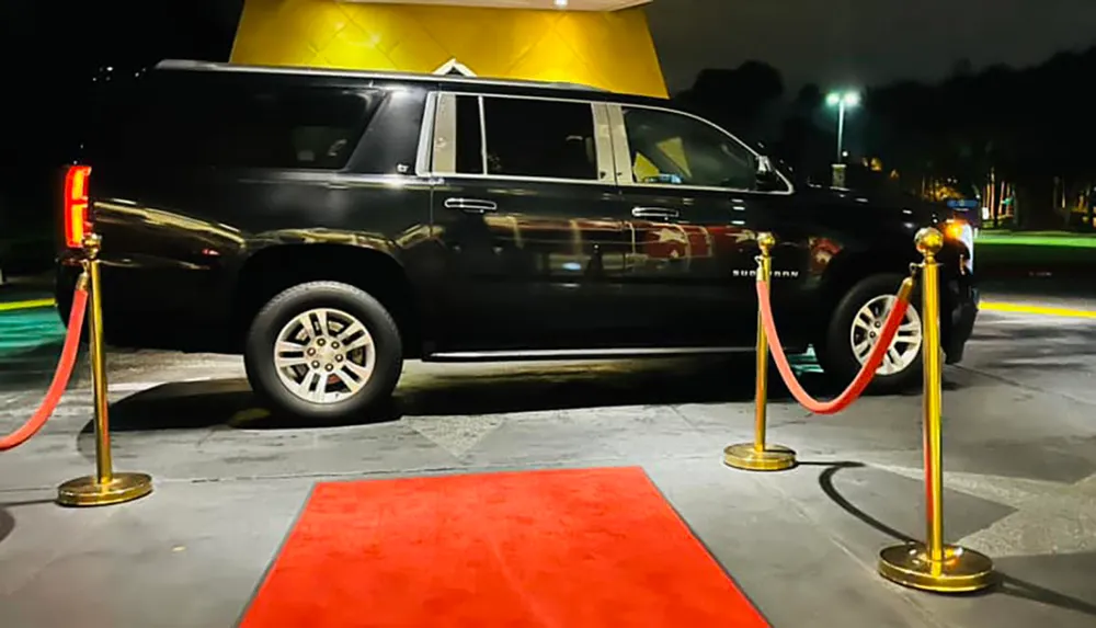 A black SUV is parked on a red carpet flanked by gold stanchions with red velvet ropes suggesting a VIP or exclusive event taking place at night