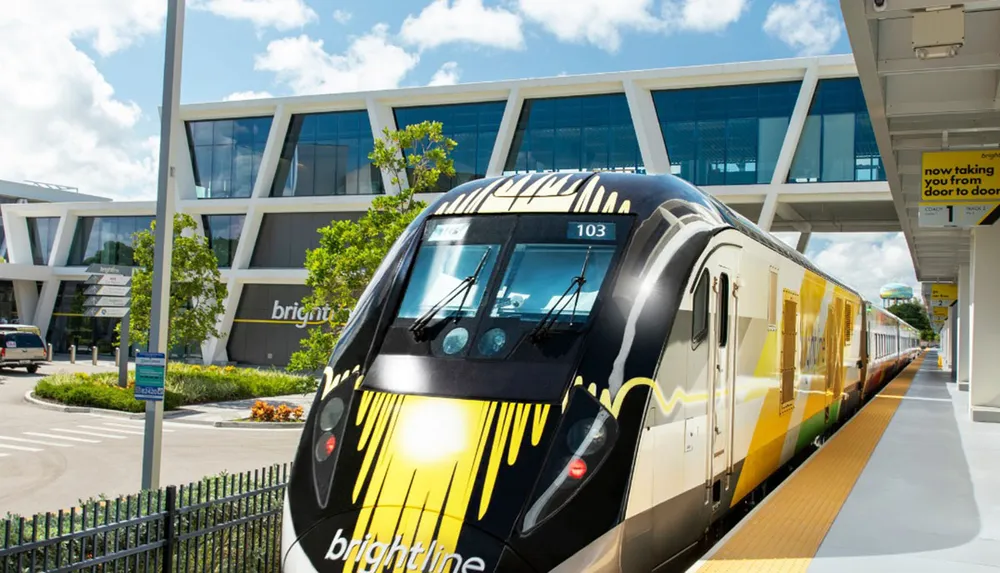 A colorful Brightline train is parked at a station on a sunny day with modern architecture in the background