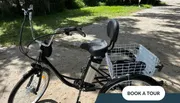 The image shows an electric bicycle with a rear cargo basket parked on a leafy path, with a button overlaid on the photo that suggests viewers can book a tour.