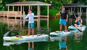 Three individuals are enjoying paddleboarding on a calm waterway, with lush greenery in the background and a dock to one side.