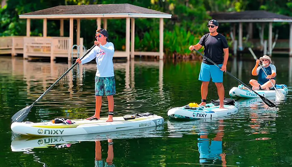 Three individuals are enjoying paddleboarding on a calm waterway with lush greenery in the background and a dock to one side