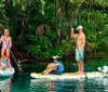 A person is standing on a paddleboard over clear water revealing underwater features with lush greenery in the background