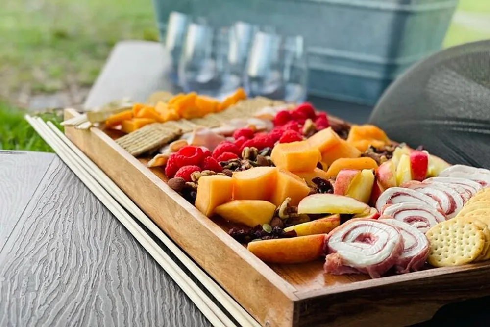 A wooden board filled with an assortment of snacks including cheeses fruits crackers and cold cuts is presented on a table outdoors suggesting a casual dining setting or picnic