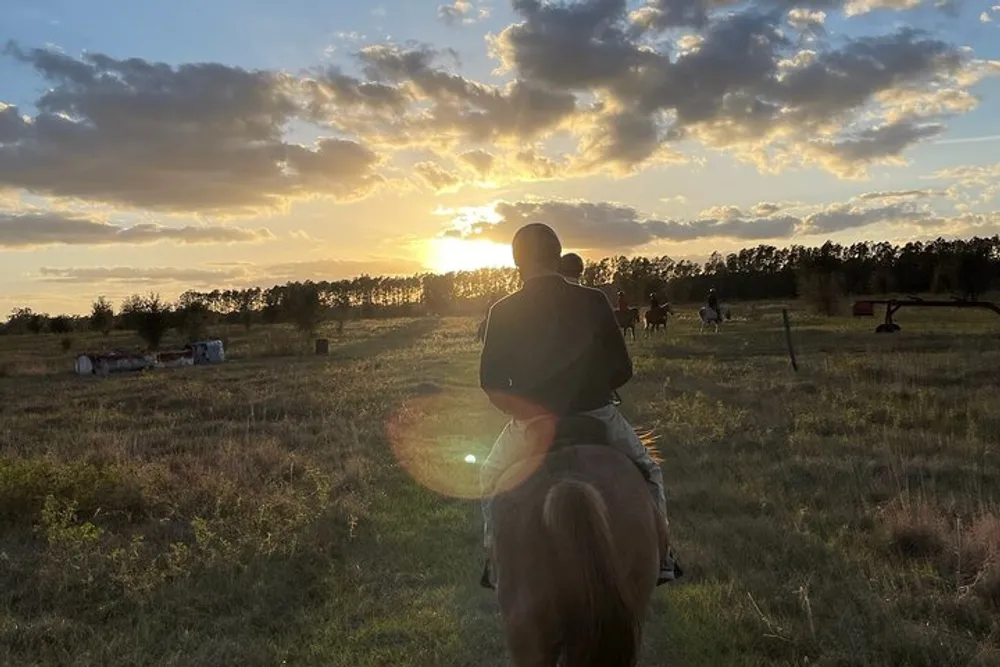 A person is riding a horse in a field at sunset with other riders in the distance and the sun peeking through scattered clouds