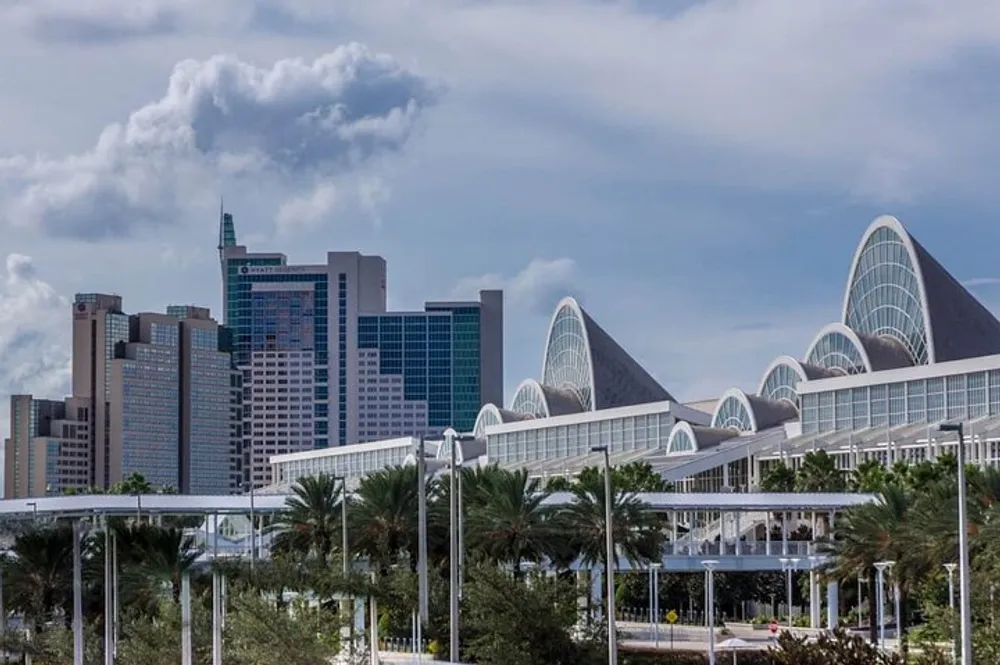 The image shows a cityscape with modern buildings and distinctive curved architecture palm trees in the foreground and a cloudy sky above