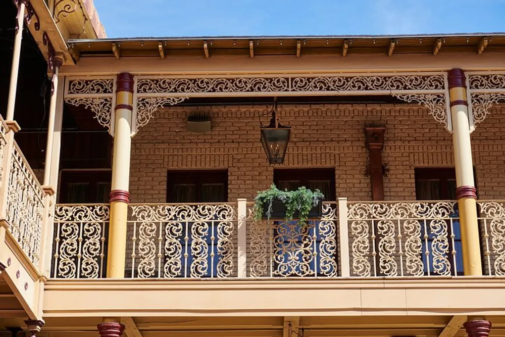 The image shows the ornate balcony of a vintage brick building with intricate iron railing and decorative woodwork under a clear blue sky