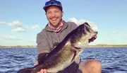 A smiling person proudly holds a large fish with a lake and a clear sky in the background.