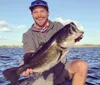 A smiling person is proudly holding a large fish on a boat with a body of water and a partly cloudy sky in the background