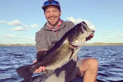 A smiling person is proudly holding a large fish on a boat, with a body of water and a partly cloudy sky in the background.