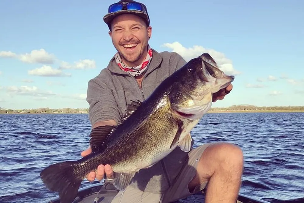 A smiling person is proudly holding a large fish on a boat with a body of water and a partly cloudy sky in the background