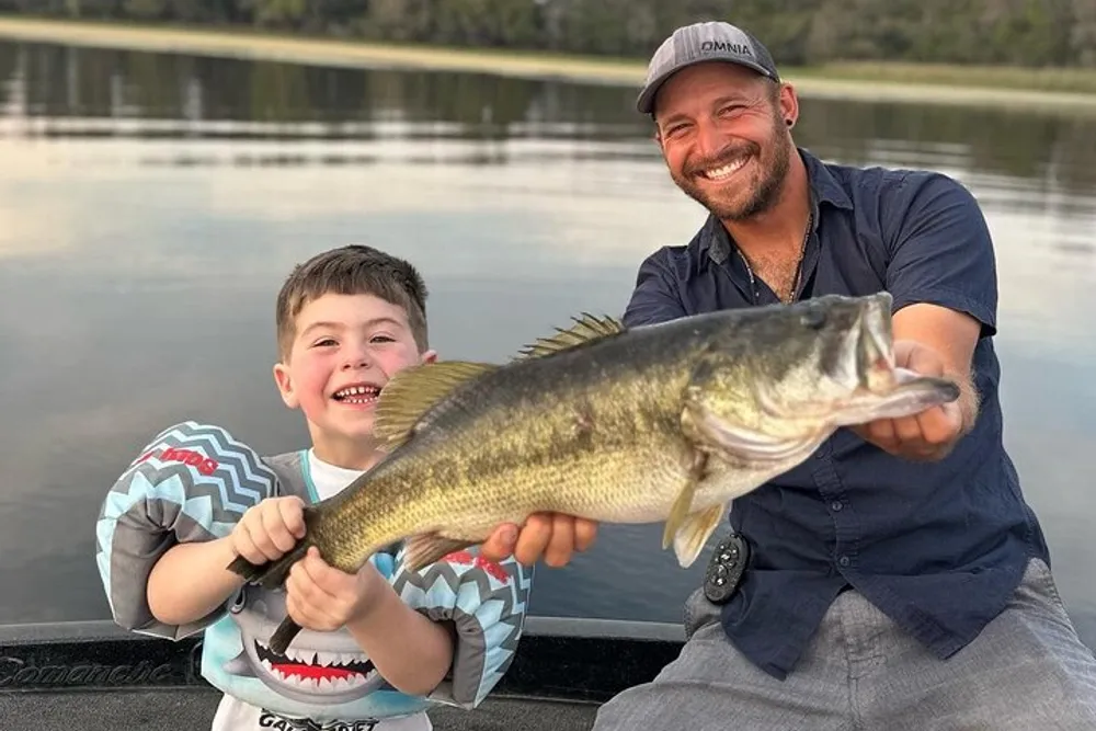 A smiling boy and an adult man are proudly displaying a large fish they caught while sitting in a boat on a calm body of water