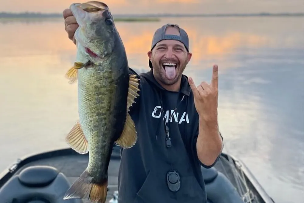 A cheerful person is holding a large fish on a boat with a water backdrop making a rock-and-roll hand gesture and sticking out their tongue