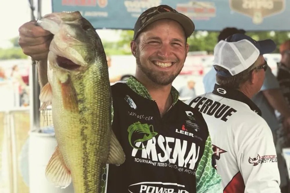 A smiling man in a fishing jersey proudly holds up a large fish hes presumably caught with a weigh-in or event backdrop behind him