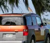 A hawk is perched on top of a parked two-tone Honda Element by a scenic lake with trees in the background