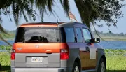 A hawk is perched on top of a parked two-tone Honda Element by a scenic lake with trees in the background.