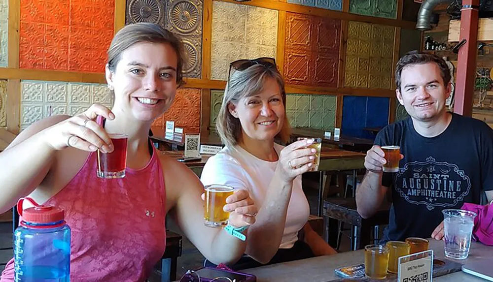 Three people are smiling and raising their glasses for a toast in a colorful casual setting