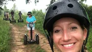 A group of people are enjoying a Segway tour on a forest trail, with a smiling woman in the foreground taking a selfie.