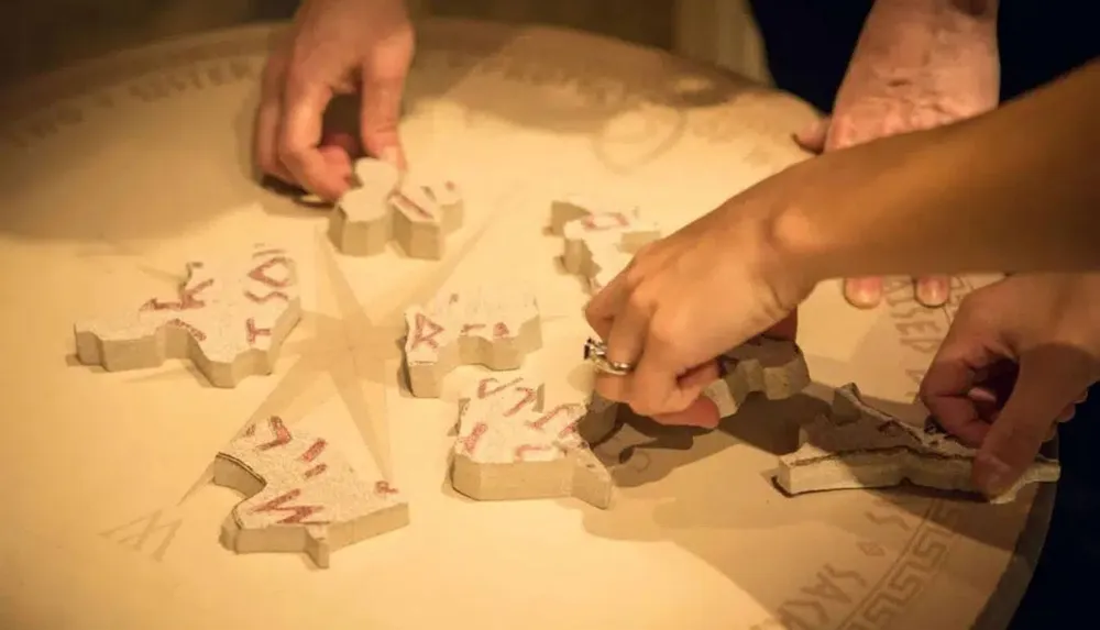 Two pairs of hands are seen assembling large puzzle-like pieces with runes on a table illuminated in warm light