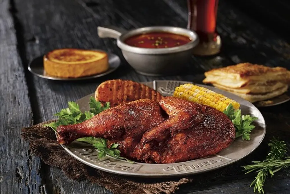 A plate with seasoned barbecue chicken legs corn on the cob and sides of sauce cornbread and a grilled slice of polenta is presented on a rustic wooden table