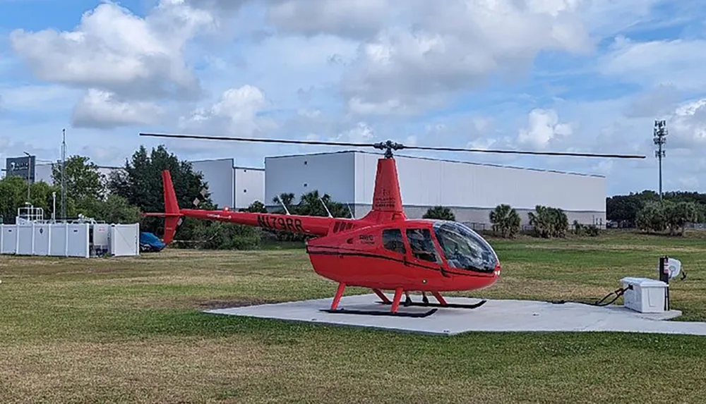 A red helicopter is parked on a landing pad in an open field with industrial buildings in the background