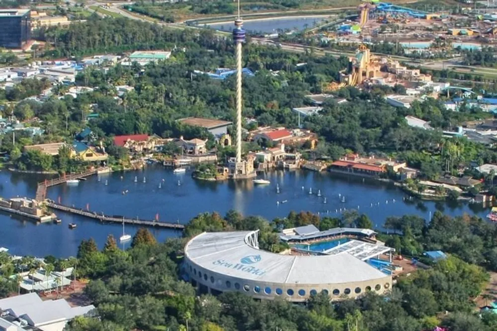 The image shows an aerial view of a theme park with a distinctive round building and a tall tower overlooking a body of water and various attractions