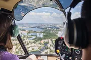 A person wearing headphones is piloting a helicopter, providing an aerial view of a landscape with water bodies and urban developments.