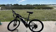 A bicycle is parked on a concrete path with a scenic backdrop of grassland and a house in the distance under a clear sky.