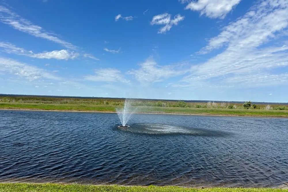 A water fountain sprays in the center of a calm pond against a backdrop of blue skies and a distant grassy field