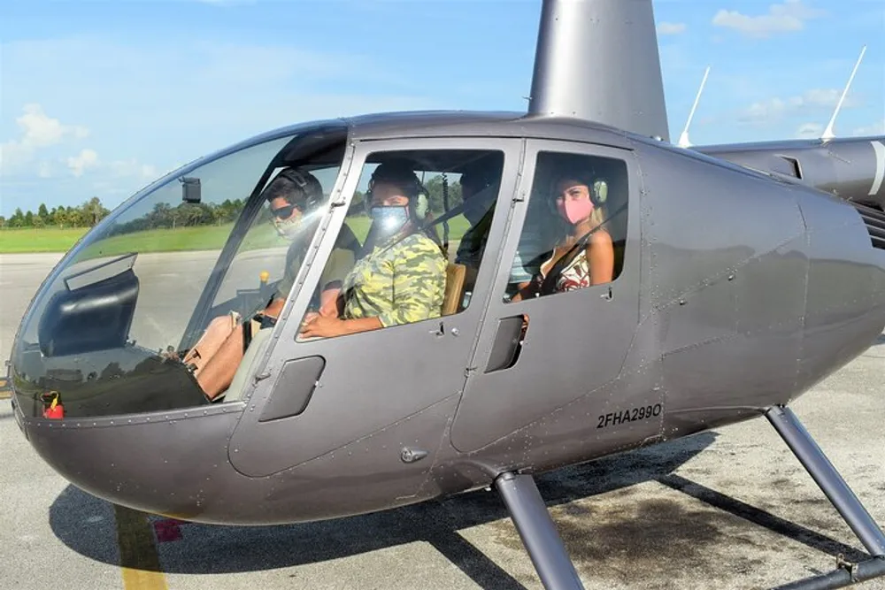 Three individuals wearing protective face masks are seated inside a helicopter which is parked on a tarmac under a clear sky