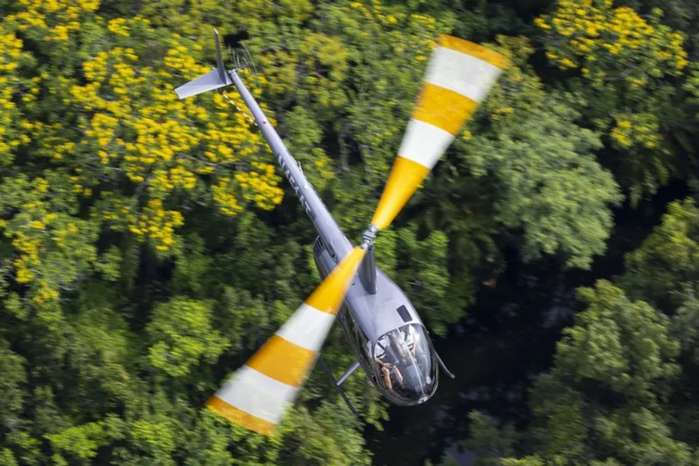 A helicopter is flying low over a dense green forest with yellow flowering trees