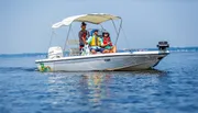 Three people wearing life jackets are enjoying a sunny day on a small motorboat with a Bimini top on a calm water body.