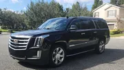 A black Cadillac Escalade SUV is parked on a suburban street in front of a house under a clear sky.