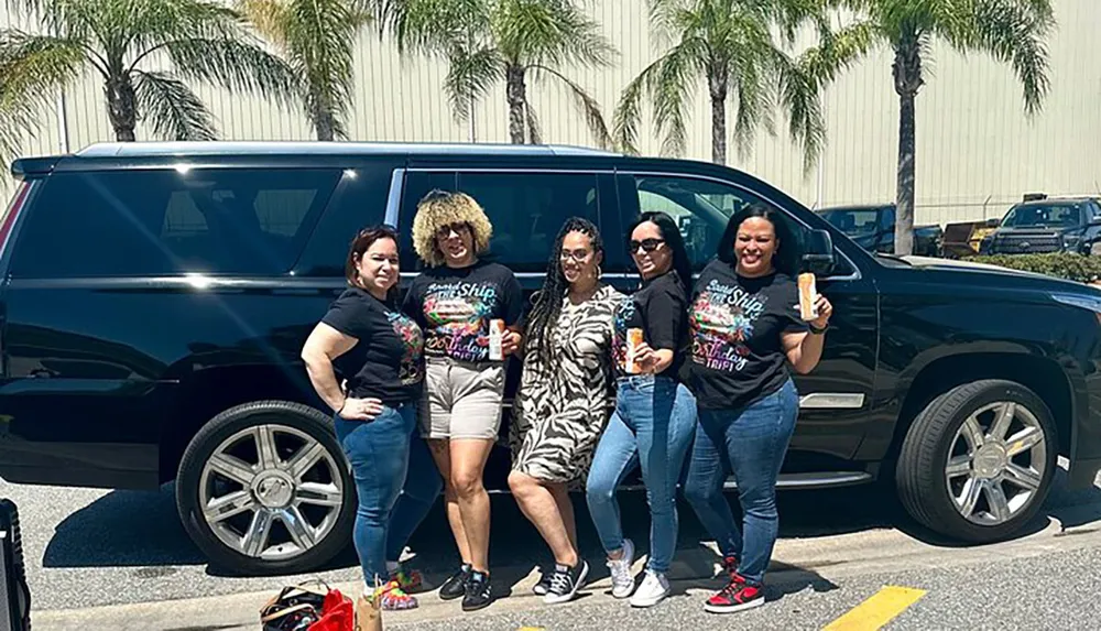 A group of five women are smiling and posing together in front of a black SUV in a sunny parking lot with palm trees in the background