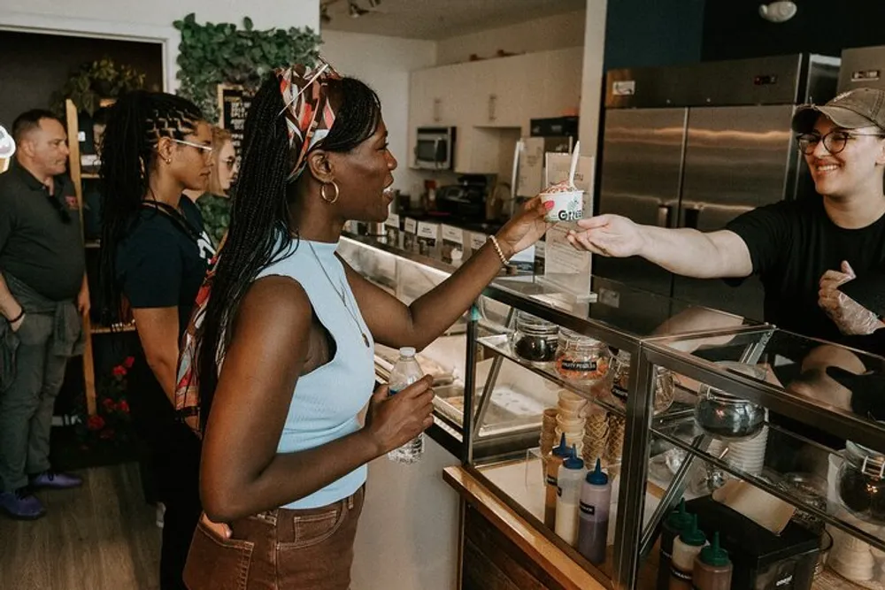 A smiling woman receives a product from a cheerful worker across a food counter with other customers waiting in line behind her