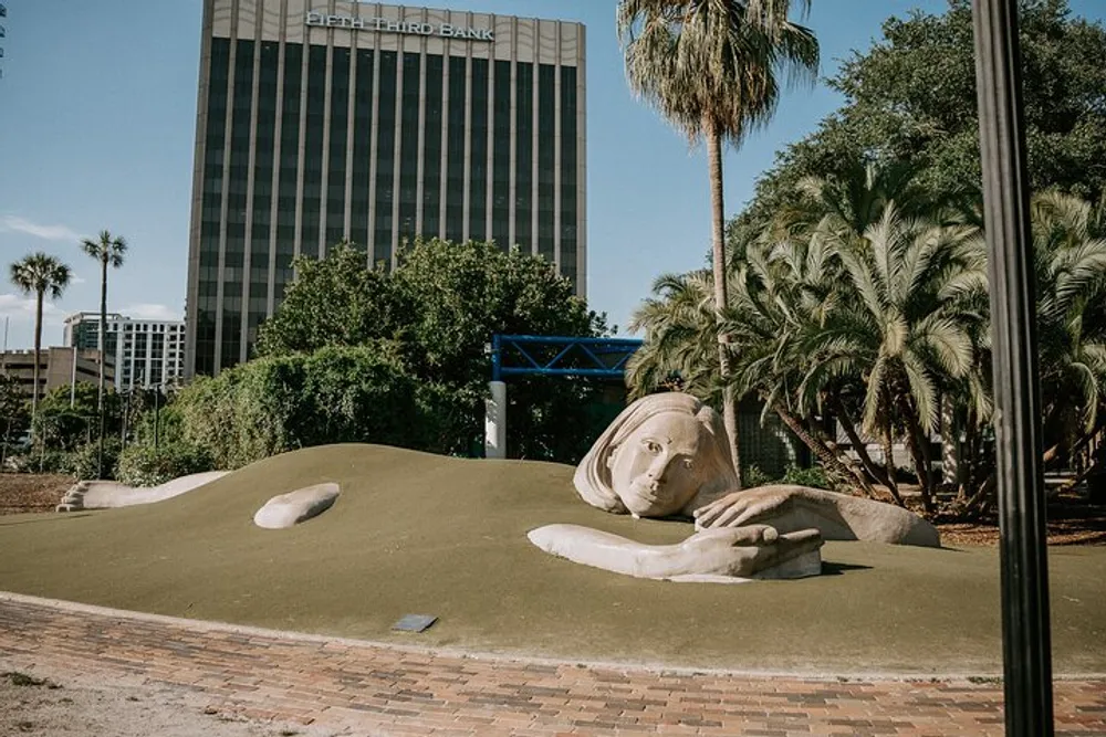 The image features an outdoor sculpture of a giant face and partial hands appearing to emerge from the ground in a park-like setting with a high-rise building in the background