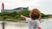 A child with curly hair is pointing towards the Space Shuttle exhibit at the Kennedy Space Center, as seen across a body of water.