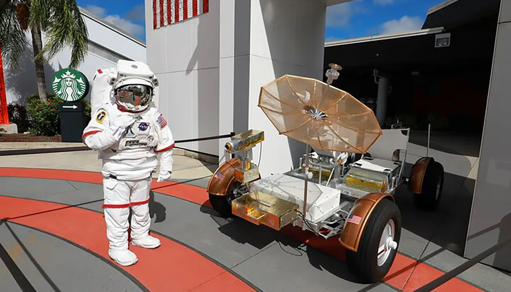 An individual in an astronaut suit is standing beside a model of a lunar rover with a Starbucks sign visible in the background