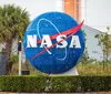 A child with curly hair is pointing towards the Space Shuttle exhibit at the Kennedy Space Center as seen across a body of water