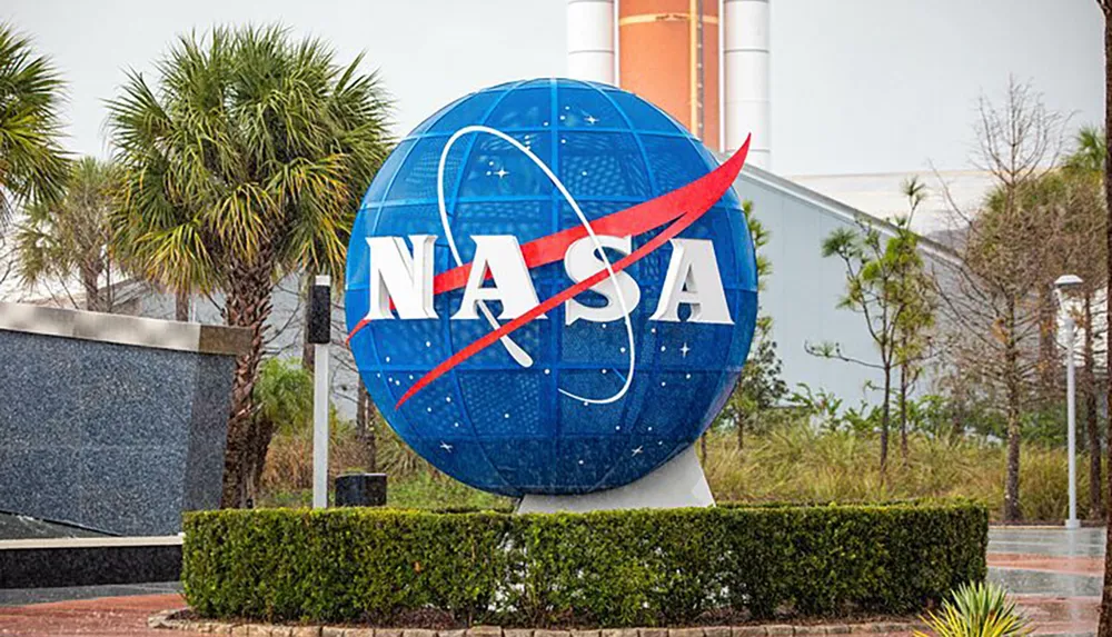 The image features a large spherical NASA sign with the agencys iconic blue and red logo in front of a landscaped area and a building with a rocket display in the background