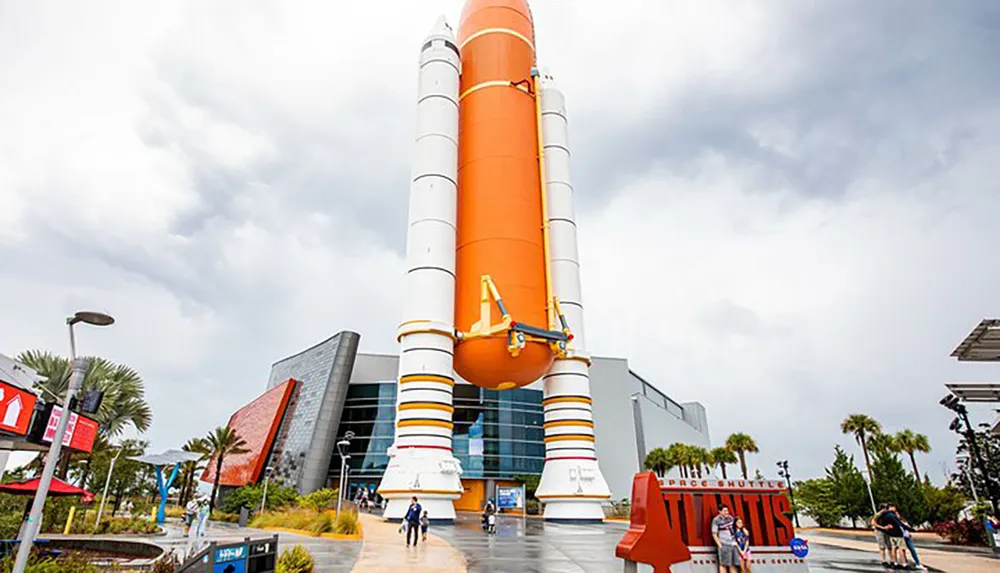 The image shows a towering orange and white space shuttle external fuel tank and solid rocket boosters on display at a space-themed visitor center with people walking around the exhibit set against a partly cloudy sky