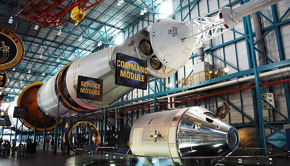 The image shows a Saturn V rocket exhibit with its stages labeled command module and service module displayed horizontally in a large hangar-like museum space